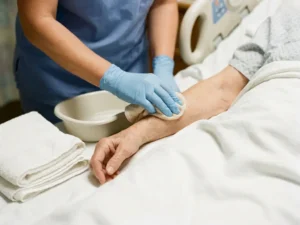 Verpleegkundige in latex handschoenen wast zachtjes de arm van een patiënt met een zeepachtige doek, witte handdoek en klinische waskom op ziekenhuisbedlinnen.