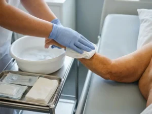 Verzorger in latex handschoenen wast voorzichtig de arm van een oudere patiënt met een doek, witte kom met zeepwater op achtergrond.