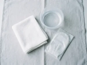 Gevouwen witte washand naast een klinische kom met helder water en een verpakte wegwerp-washandschoen op wit linnen.