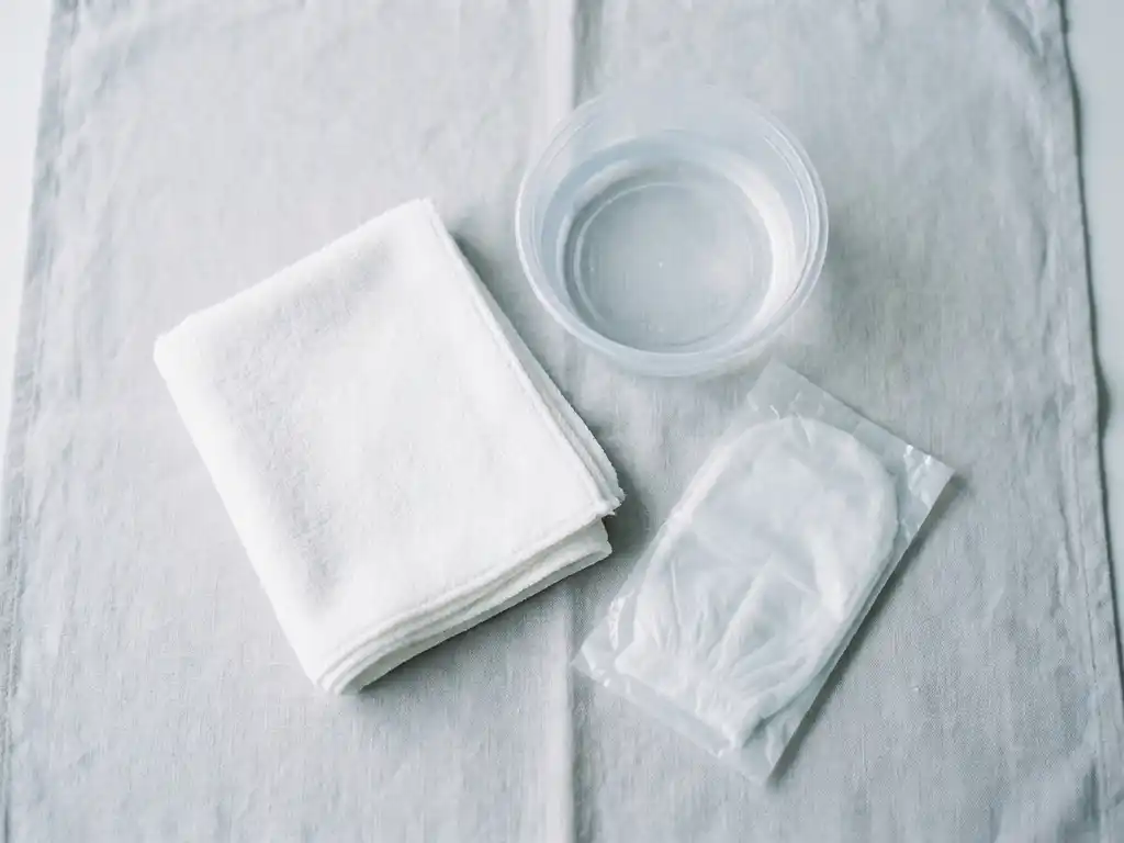 Gevouwen witte washand naast een klinische kom met helder water en een verpakte wegwerp-washandschoen op wit linnen.