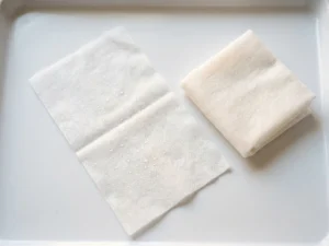 Wegwerp washandjes op wit oppervlak, één uitgevouwen met zichtbare textuur en vochtige druppels, één gevouwen ernaast.