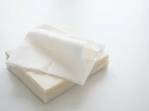 Stapel gevouwen disposable washandjes op wit oppervlak, één gedeeltelijk opengevouwen met zichtbare zachte vezelstructuur.