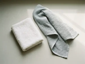 Zacht gevouwen wit washandje op klinisch oppervlak naast een uitgevouwen doek, minimalistische ziekenhuisesthetiek in witte en grijze tinten.